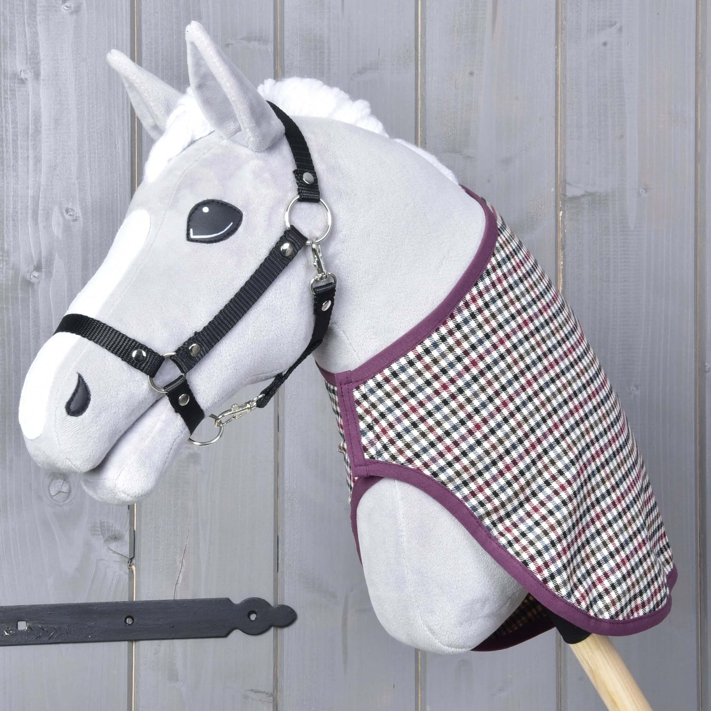 Ein graues Hobby Horse Spielzeug von Hobby Horse LarDen mit schwarzem Halfter für Hobby Horse und der Decke western Colorado, montiert auf einem Holzstab und vor einer grauen Holzwand.