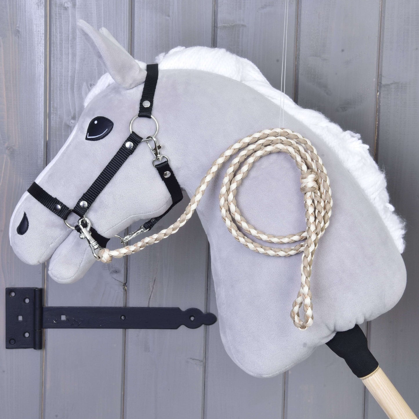 Hobby Horse - Corde d'attache tricotée Double Latte de Hobby Horse LarDen, cheval bâton gris avec bride noire et rênes tressées, présenté devant un mur en bois gris.