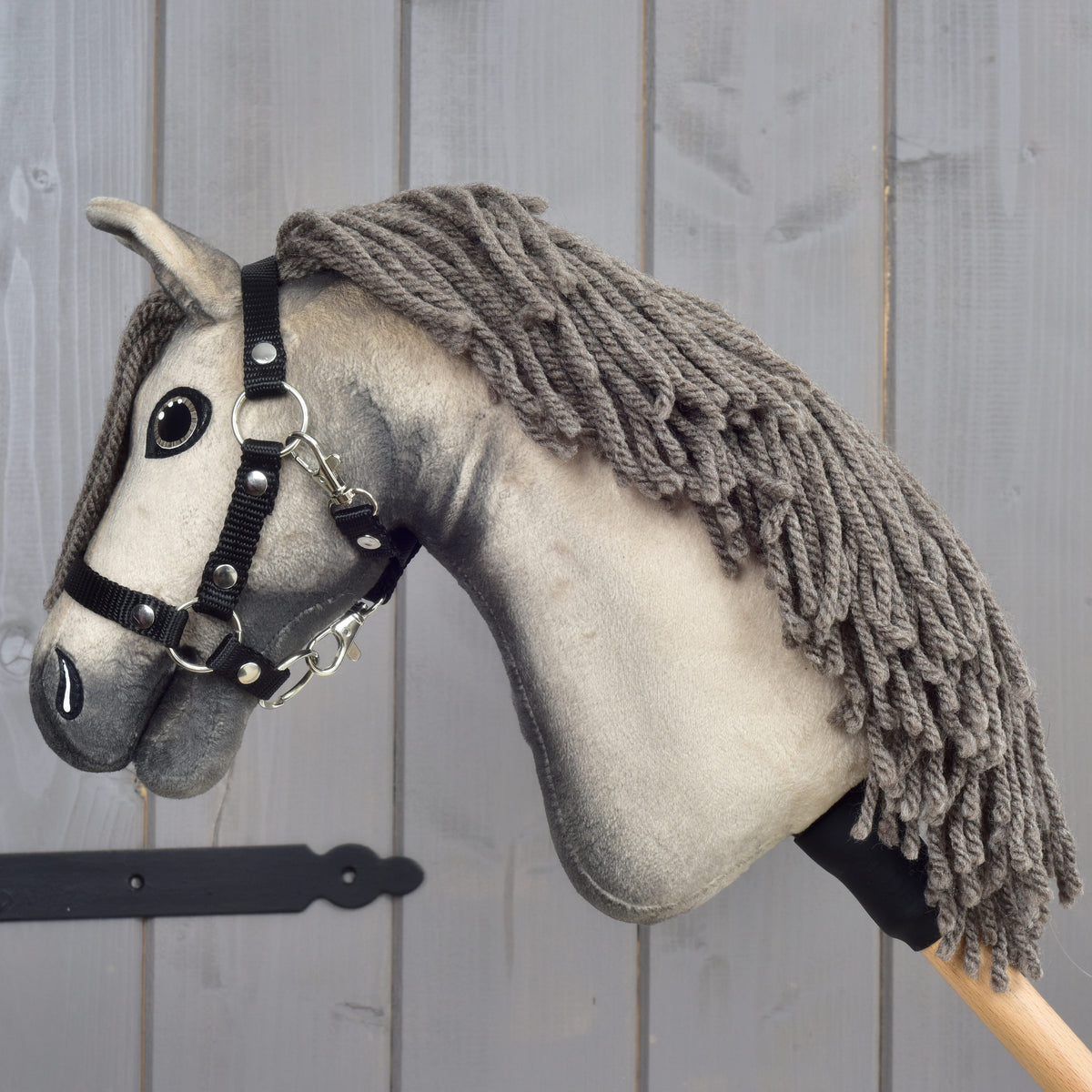 The Hobby Horse LarDen "Hobby Horse Dusty with Black Halter" (size XS), featuring a gray coat, yarn mane, and black halter, is mounted on a wooden stick and placed against a light gray wooden wall.