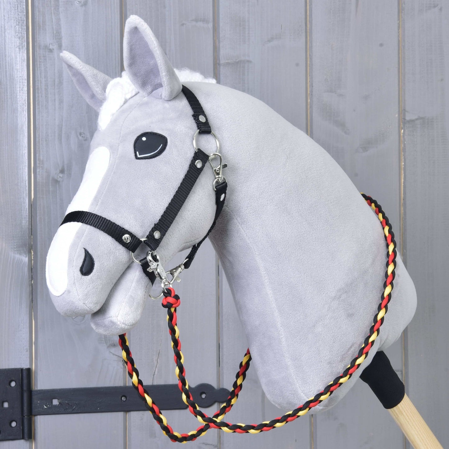 Rênes Drapeau de Hobby Horse LarDen sur un Hobby Horse gris en peluche monté sur un bâton en bois, avec harnais noir et rênes faites main en corde de polypropylène tressée rouge, jaune et noire, devant un mur en bois.