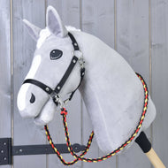 Hobby Horse LarDen Zügel Flag ist ein graues Plüsch-Hobby Horse auf einem Holzstab mit schwarzem Geschirr und handgefertigten Zügeln aus geflochtenem Polypropylenseil in Rot, Gelb und Schwarz, gezeigt vor einer Holzwand.