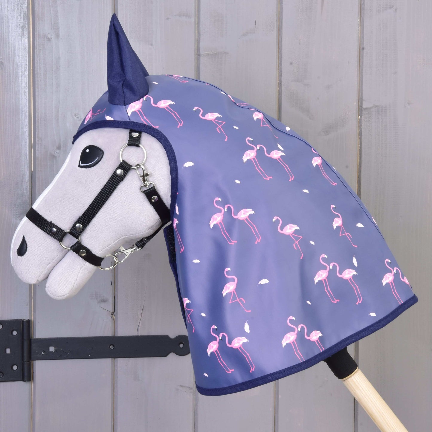 Hobby Horse gris de Hobby Horse LarDen avec bride noire, portant l’Imperméable Flamant Rose, une capuche bleue pour tous les temps avec flamants roses et points blancs, sur fond en bois.