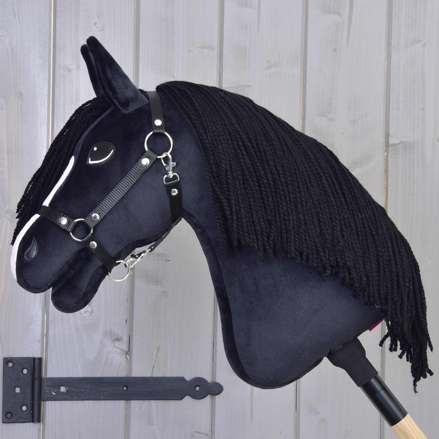 Hobby Horse Freya mit Schwarzem Halfter von Hobby Horse LarDen mit weißer Blesse und schwarzer Garnmähne auf einem Holzstab, vor einer hellgrauen Holzwand – perfekt für jeden Pferdefan in der Familie.