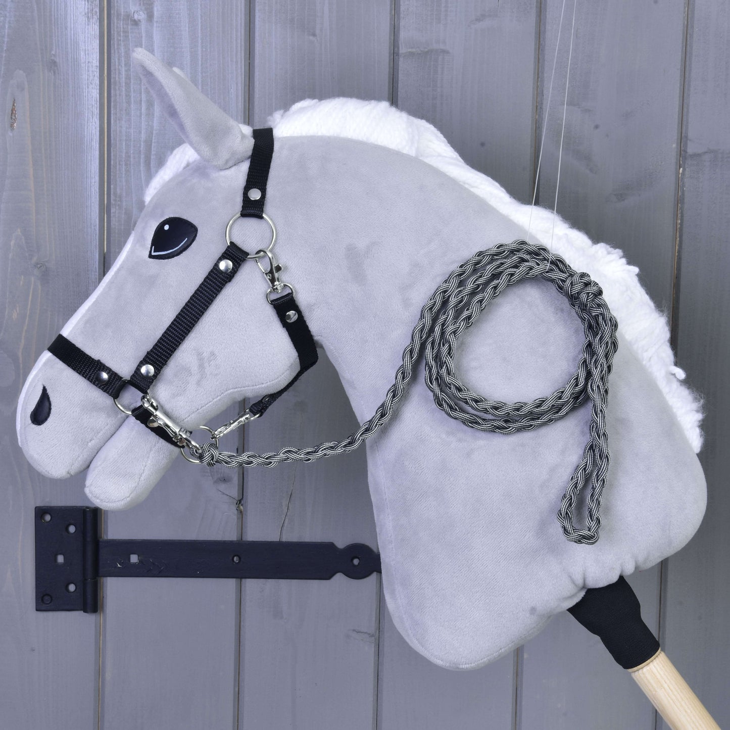 Hobby Horse - Corde d'attache tricotée Graphite de Hobby Horse LarDen, tête en peluche grise avec bride noire et longe graphite tricotée à la main sur un bâton en bois, présentée devant un mur en bois gris.