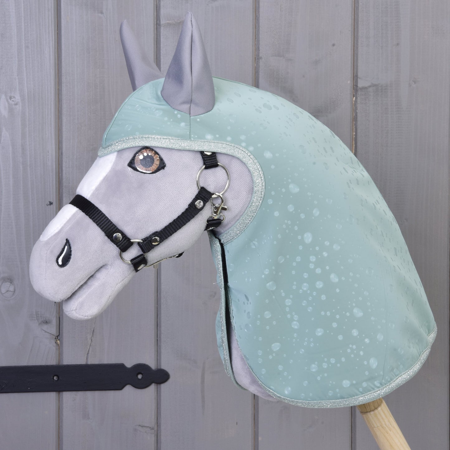 Hobby Horse LarDen « Imperméable Greeny », cheval en peluche vêtu d’un imperméable vert à motif gouttes d’eau et d’une bride noire, debout près d’un mur en bois gris. Les détails brodés et les oreilles dressées apportent un côté ludique pour jouer par tous les temps.