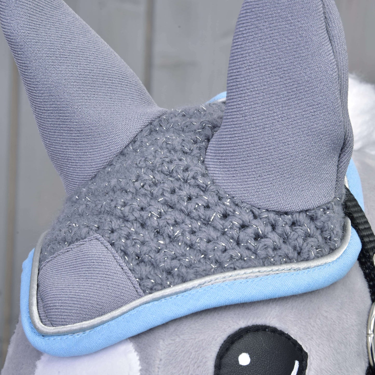 Nahaufnahme eines Plüschtiers von Hobby Horse LarDen mit „Ohrenkappe gehäkelt grau“, mit zwei grauen gehäkelten Ohren, einem gestrickten grauen Einsatz mit Silberfäden, hellblauer Einfassung und einem Teil eines schwarz-weißen Auges. Unscharfer Hintergrund.