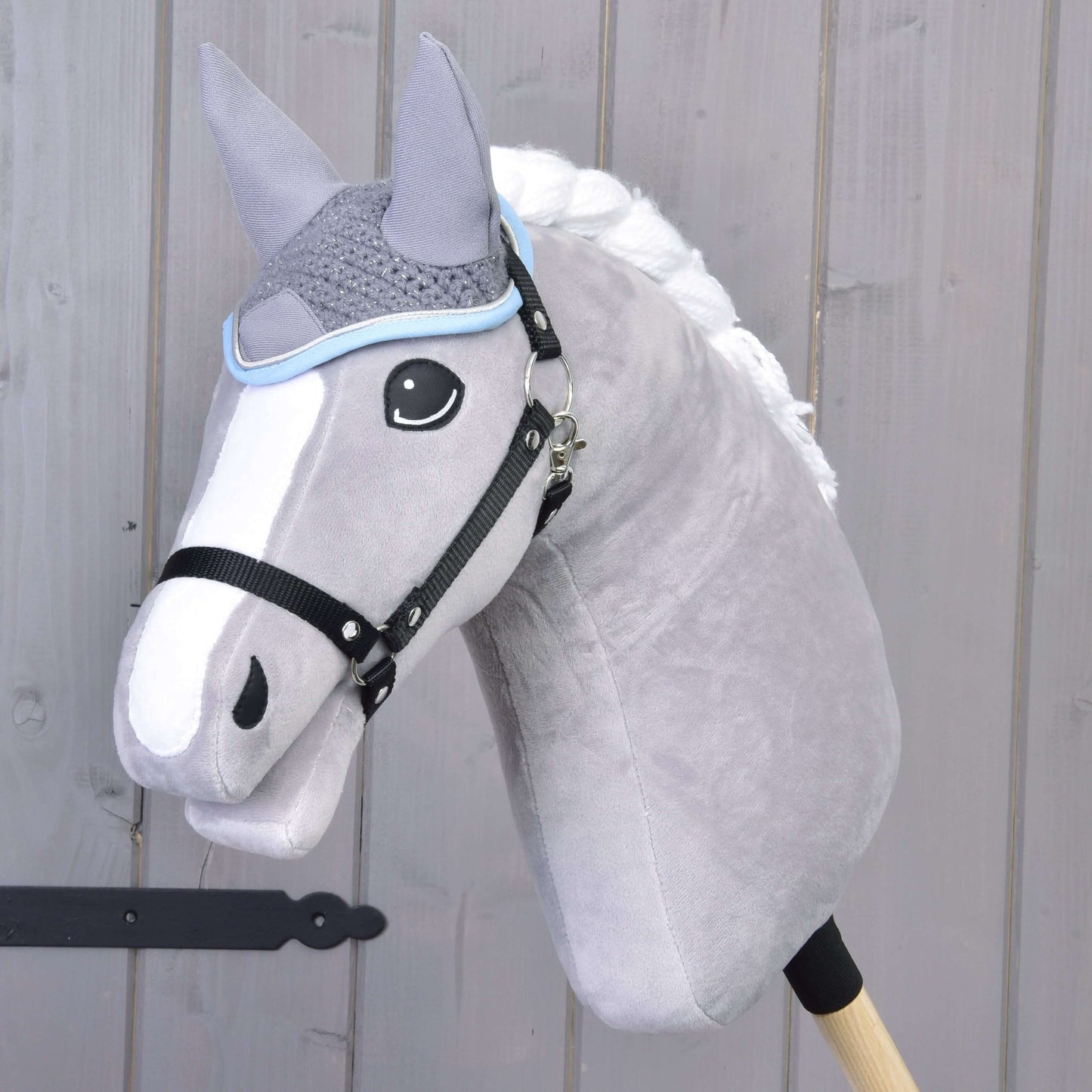 Ohrenkappe gehäkelt grau von Hobby Horse LarDen auf einem plüschigen grauen Hobby Horse mit weißer Mähne und schwarzem Zaumzeug, auf einem Holzstab vor einer hellen Holzwand.