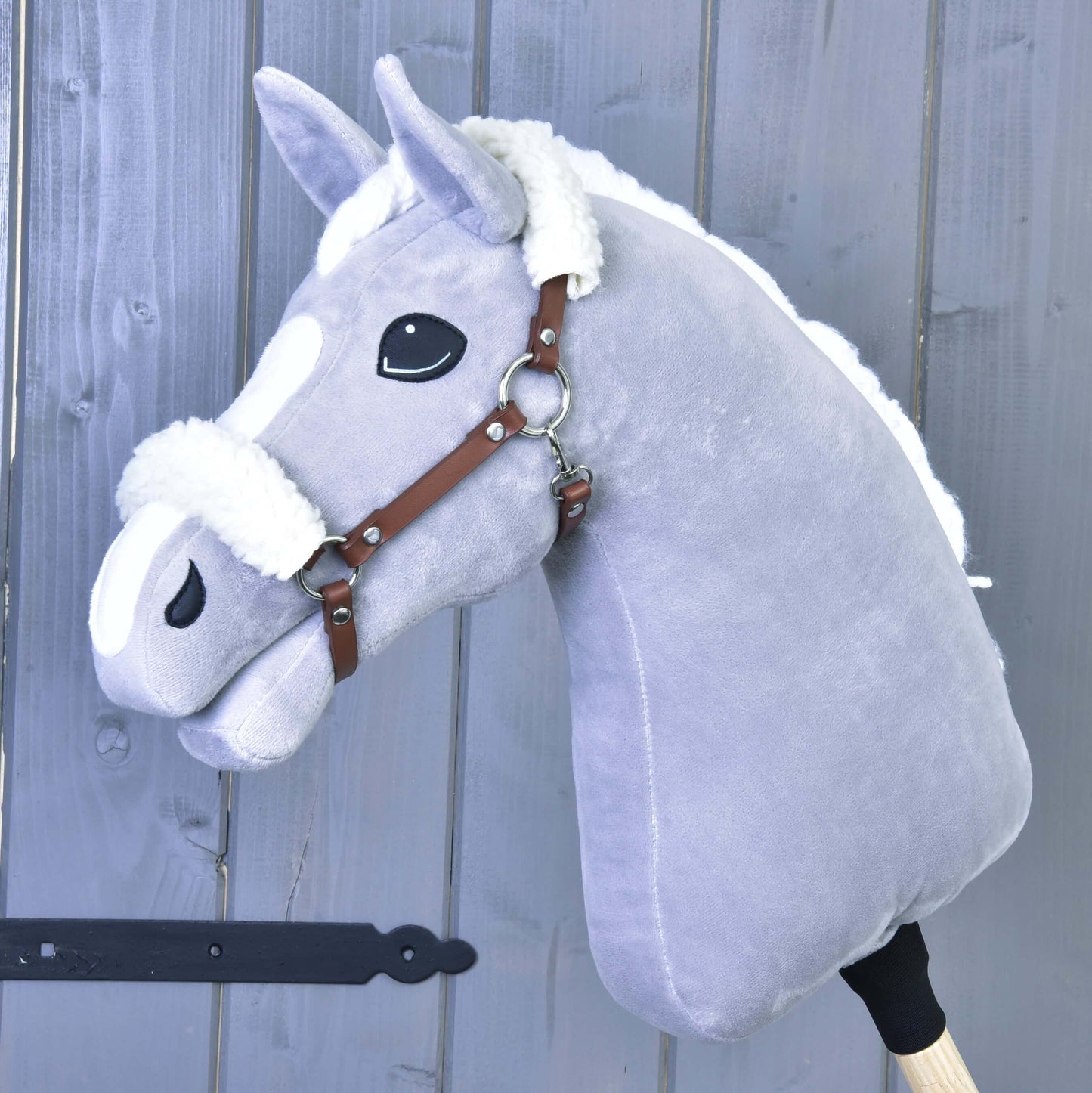 Das graue Plüsch-Hobby Horse von Hobby Horse LarDen mit weißer Mähne und Braunes Halfter mit Fell - Kunstleder ist auf einem Holzstab montiert und vor einer grauen Holzwand dargestellt.
