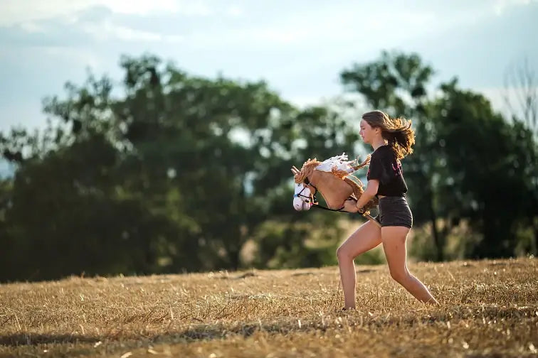 Eine junge Person läuft beim Hobby Horsing mit einem Spielzeug-Hobby Horse durch ein trockenes, grasbewachsenes Feld, mit Bäumen und blauem Himmel im Hintergrund.