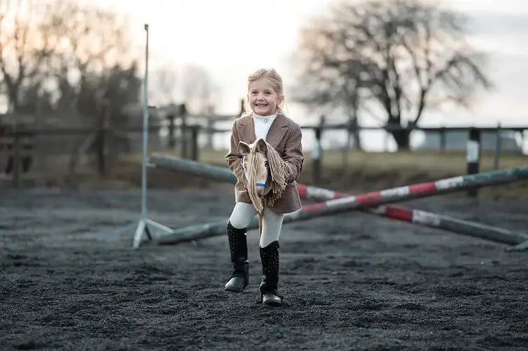 Ein junges Mädchen lächelt beim Hobby Horsing und tut so, als würde sie draußen auf einem Spielzeug-Hobby Horse auf einem Sandplatz mit gekreuzten Stangen und Bäumen im Hintergrund reiten. Sie trägt eine braune Jacke, weiße Hose und schwarze Reitstiefel.
