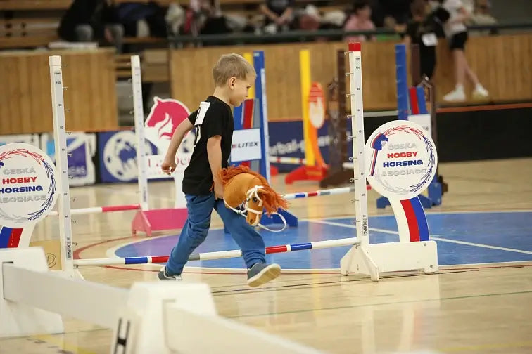 Ein kleiner Junge nimmt an einem Hobby Horse-Wettbewerb teil und läuft mit einem Spielzeugpferd über den Hallenboden, umgeben von bunten Sprunghindernissen und Schildern einer Hobby Horsing-Veranstaltung.