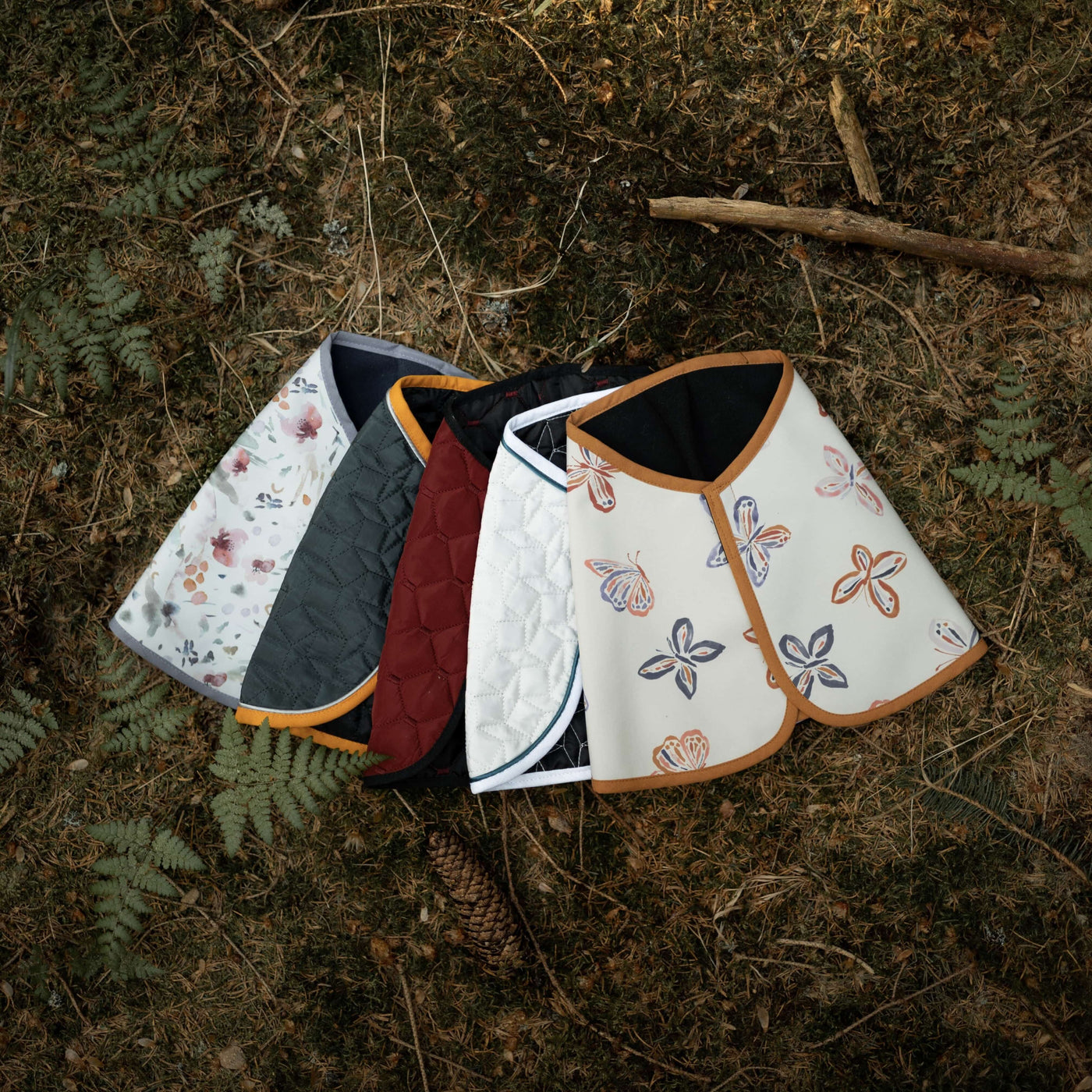 Fünf gesteppte, capeartige Kleidungsstücke mit Blumen-, einfarbig dunklem, weißem und Schmetterlingsdesign liegen fächerförmig auf einem Waldboden mit trockenen Blättern und Farnen, als würden sie in der Natur auf die nächste Runde Hobby Horsing warten.