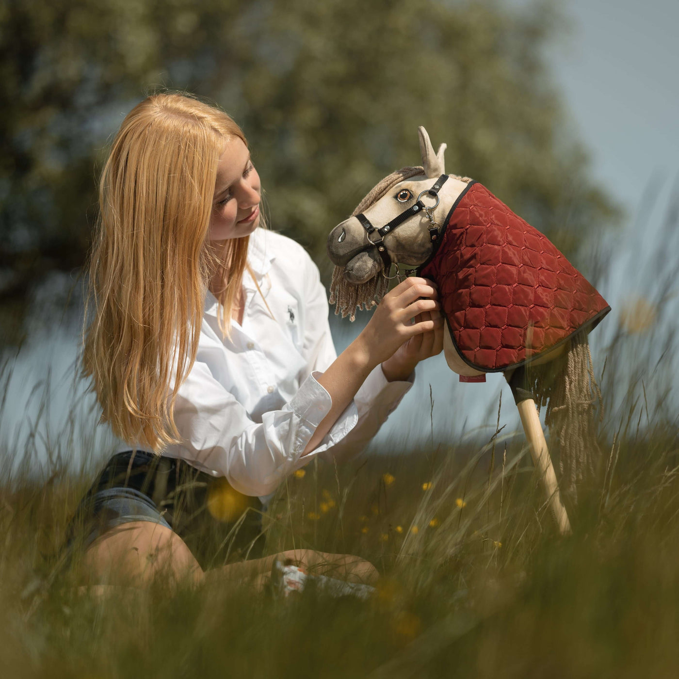 Eine junge Frau mit blonden Haaren sitzt im hohen Gras und lächelt, während sie ihren Begleiter fürs Hobby Horsing hält und ansieht – ein Hobby Horse mit rotem gestepptem Mantel. Bäume und verschwommenes Grün füllen den Hintergrund.