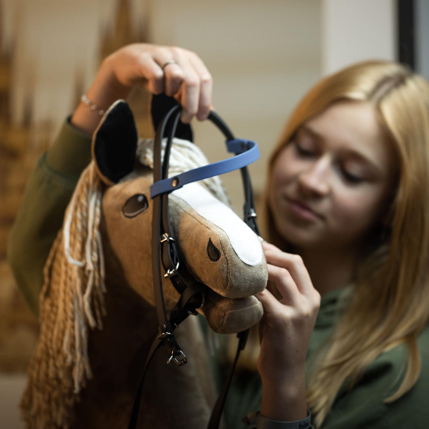 Ein Mädchen mit blonden Haaren legt ihrem Hobby Horse ein Zaumzeug an und konzentriert sich sorgfältig auf das Anpassen der Riemen – das fängt den Geist von Hobby Horsing ein.