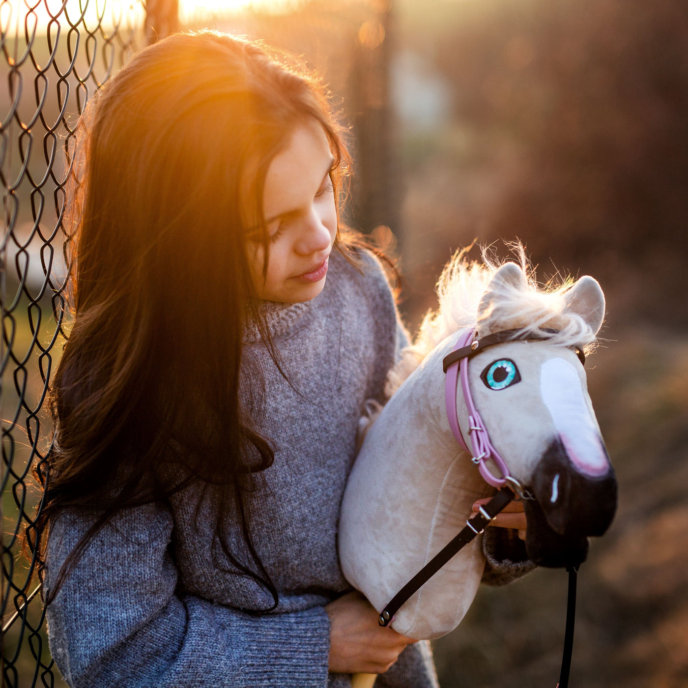 Ein junges Mädchen mit langen dunklen Haaren und grauem Pullover steht bei Sonnenuntergang an einem Drahtzaun und hält sein geliebtes Hobby Horse – eine Anspielung auf die verspielte Welt des Hobby Horsing –, dessen weiße Mähne und rosa Zaumzeug im goldenen Licht leuchten.