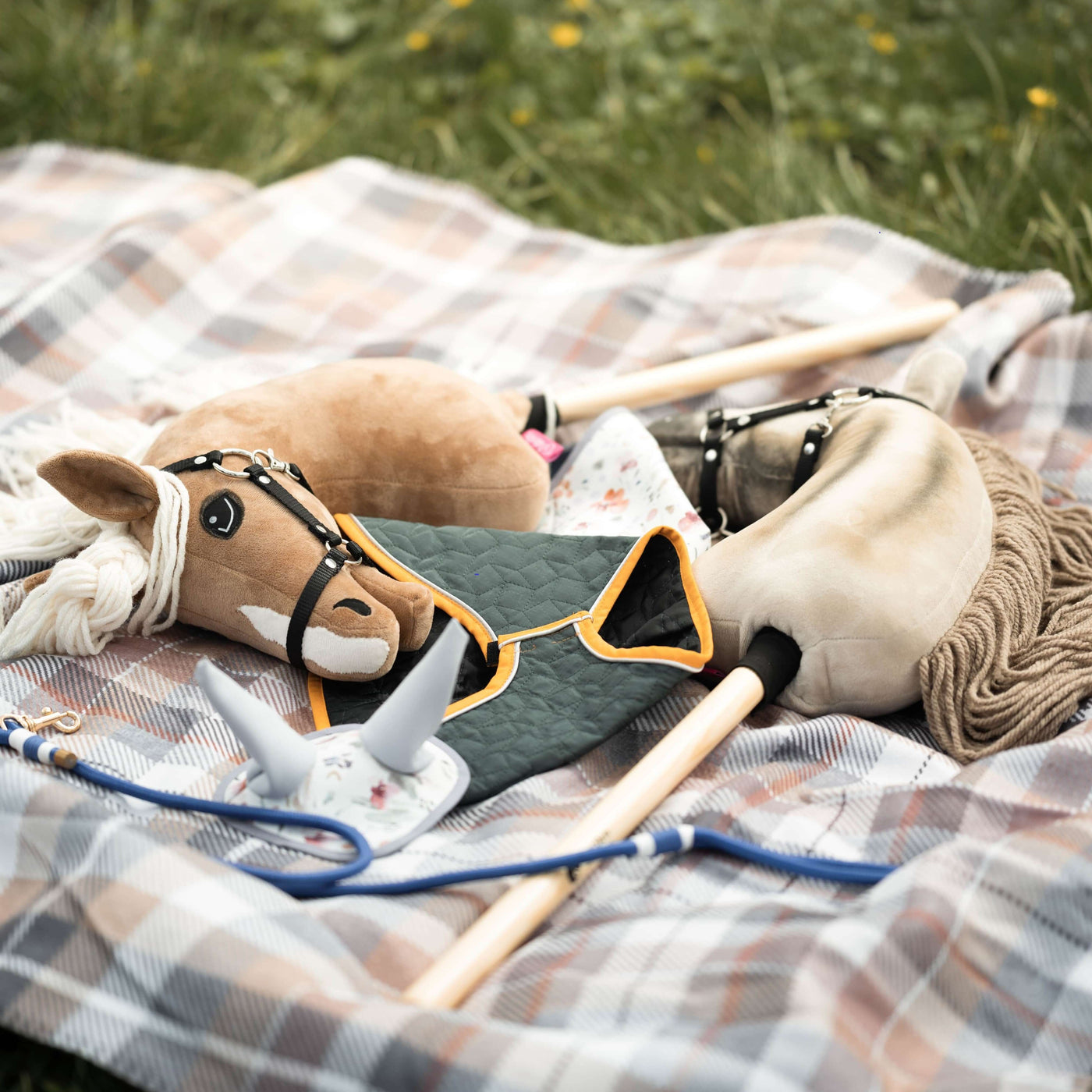 Zwei Hobby Horse Spielzeuge mit Stoffköpfen, Zügeln und Holzstöcken liegen auf einer karierten Decke auf dem Gras, zusammen mit einem kleinen grauen Spielzeug, das einem Pferdekopf ähnelt, und einem grünen gesteppten Tuch – perfekt für fantasievolle Hobby Horsing Abenteuer.