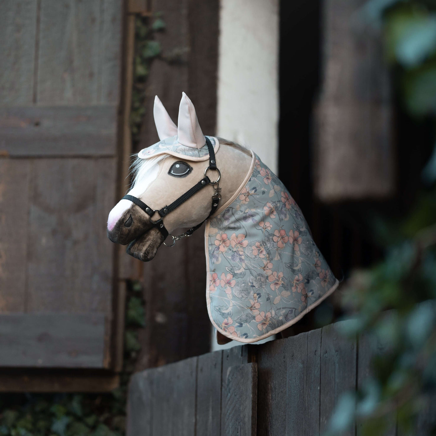 Ein realistisches Hobby Horse, perfekt für Hobby Horsing, mit Zaumzeug und blauer Decke mit Blumenmuster liegt über einem dunklen Holzzaun, vor Holzläden und üppigem Grün.