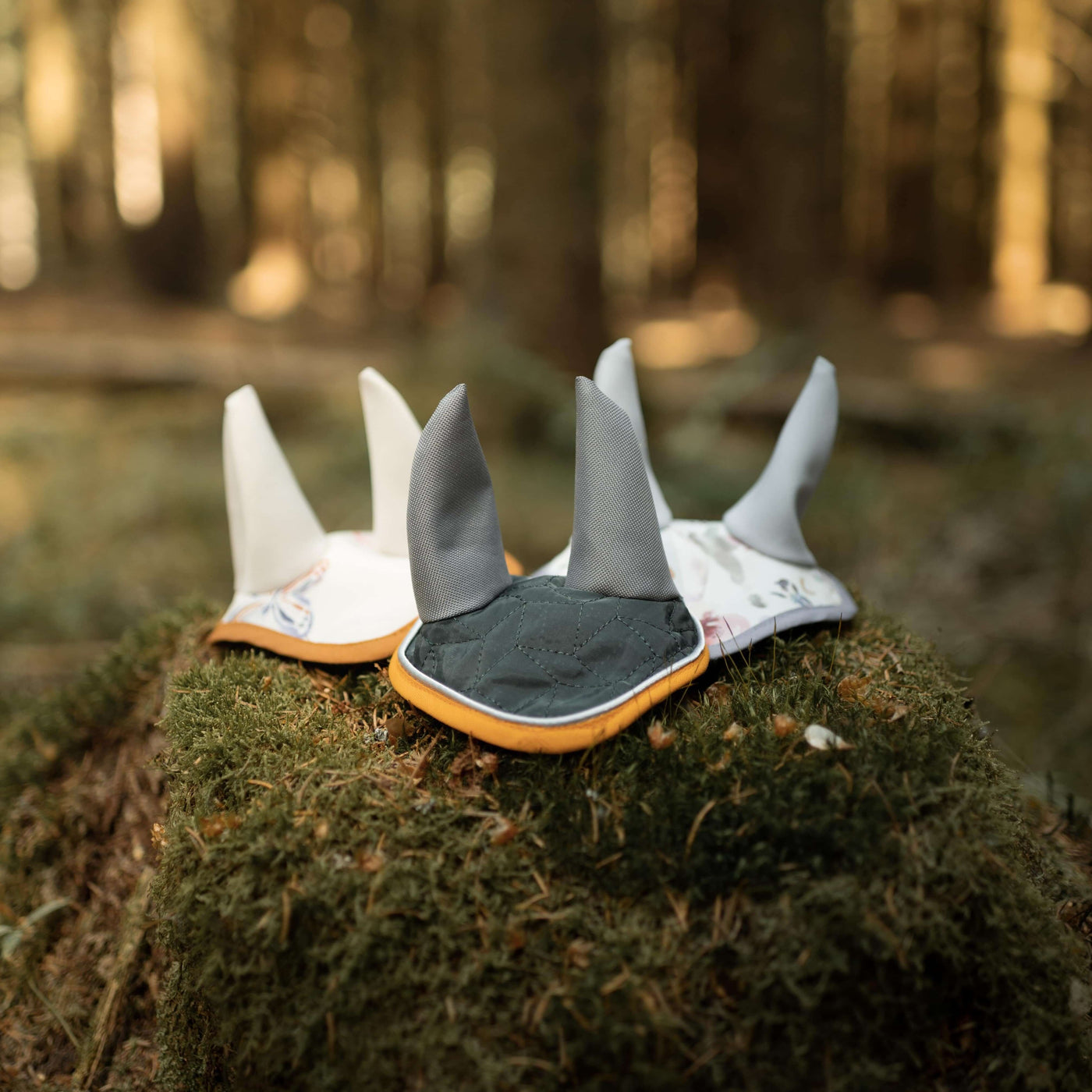 Drei weiche Babyschuhe mit Tierohren-Design liegen auf einem moosbedeckten Baumstumpf in einem sonnigen Wald und erinnern an den verspielten Geist des Hobby Horsing. Die Hausschuhe in Weiß, Grau und mit Muster haben jeweils charmante spitze Ohren.