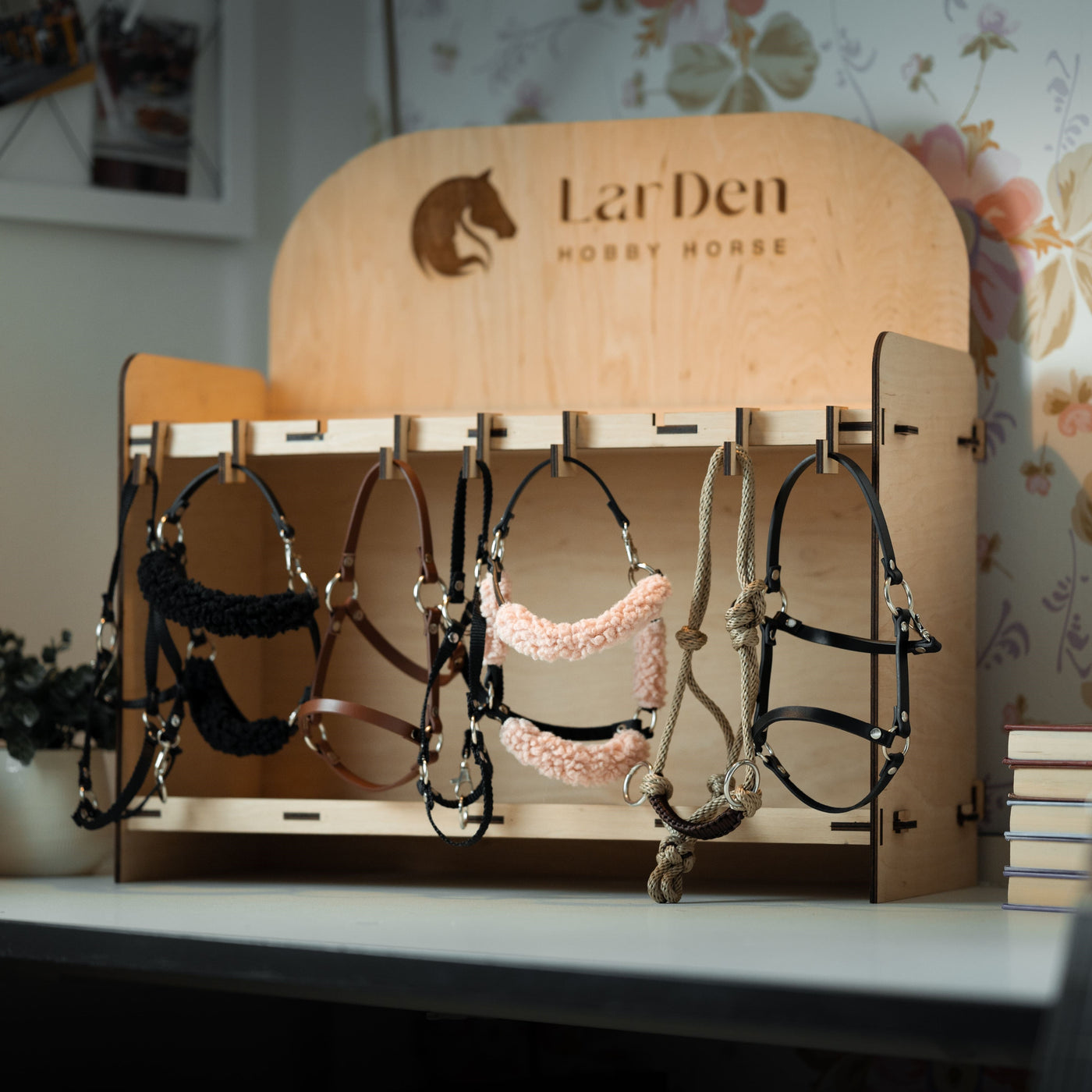 Ein hölzerner Präsentationsständer mit der Aufschrift LarDen Hobby Horse hält mehrere farbenfrohe Zaumzeuge und Halfter für Hobby Horse – perfekt für Hobby Horsing-Fans. Der Ständer steht auf einem weißen Tisch neben einem Bücherstapel, mit floraler Tapete im Hintergrund.