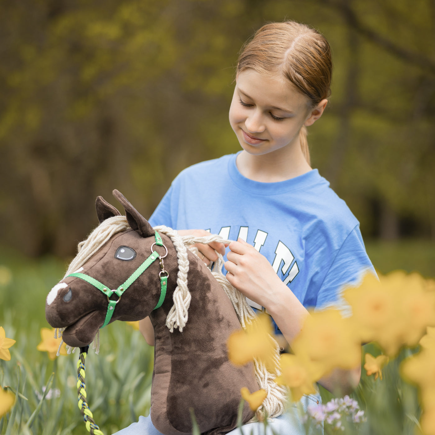 Ein Mädchen in einem blauen Shirt lächelt, während es sein braunes Hobby Horse hält und streichelt, und in einem Feld mit gelben Narzissen sitzt, mit unscharfen grünen Bäumen im Hintergrund.
