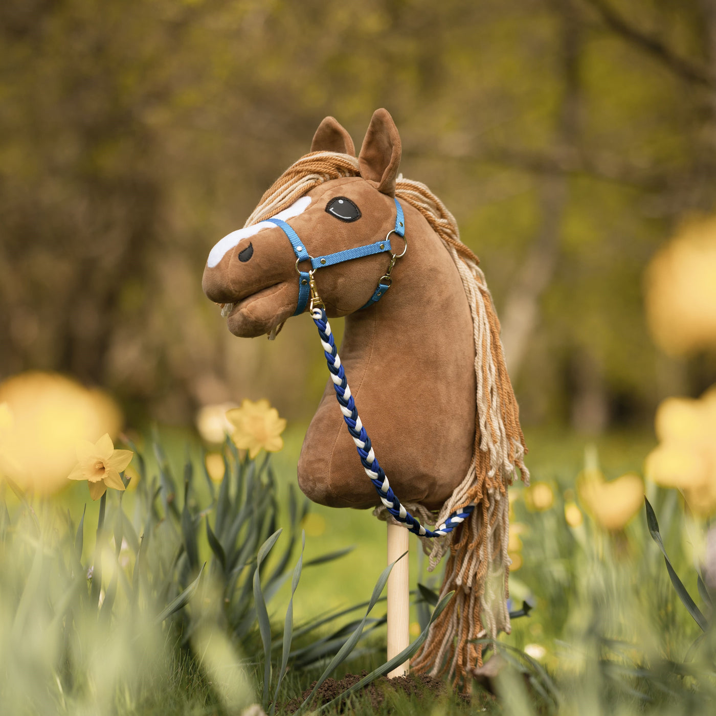 Ein braunes Hobby Horse mit blauem Zaumzeug und Wollmähne steht zwischen gelben Narzissen in einem grasbewachsenen, sonnigen Garten – eine perfekte Szene für Hobby Horsing vor verschwommenen grünen Bäumen im Hintergrund.