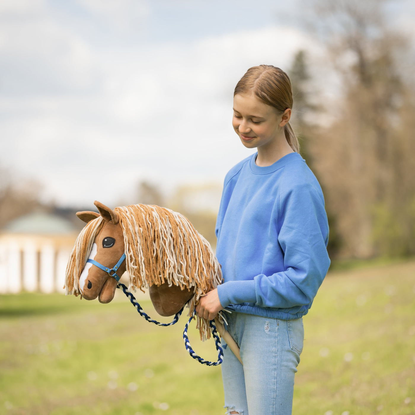 Eine junge Person in blauem Sweatshirt und Jeans steht draußen und lächelt auf ein braunes Hobby Horse mit Seilzaumzeug hinab und genießt den Spaß am Hobby Horsing auf einer Rasenfläche mit Bäumen im Hintergrund.