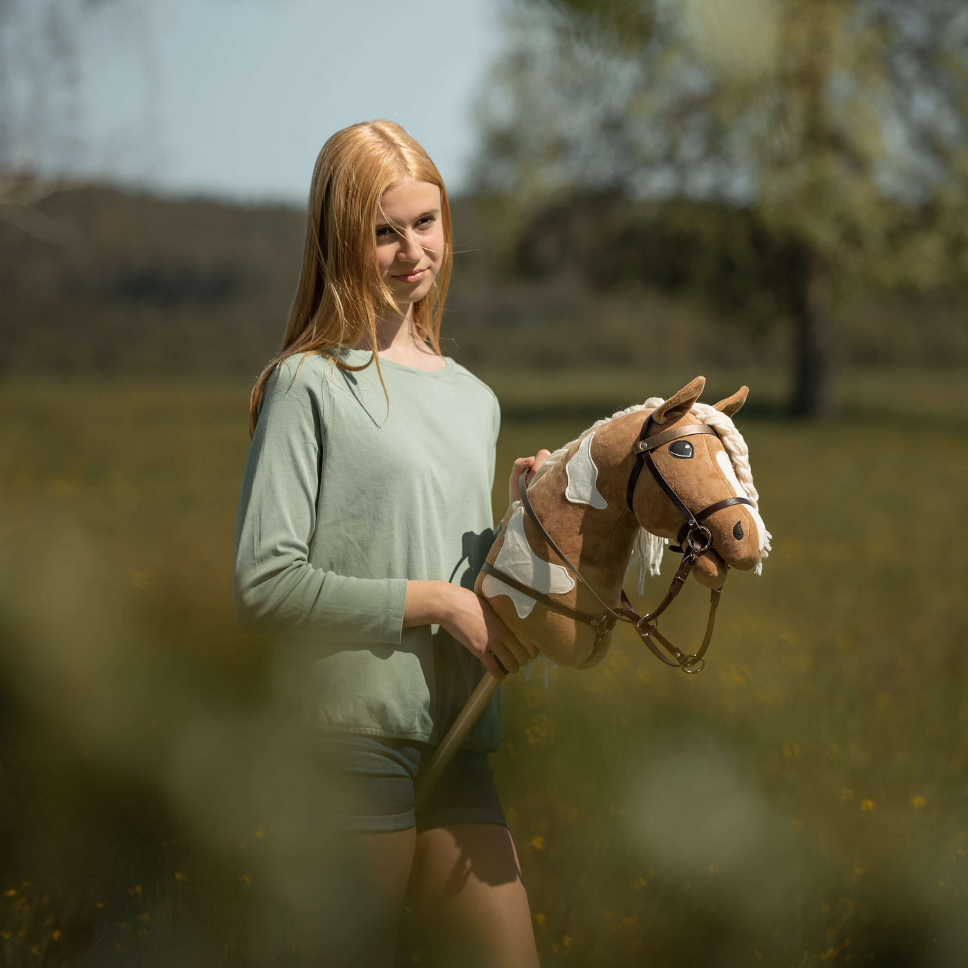 Ein junges Mädchen mit langen blonden Haaren steht draußen auf einem grünen Feld und hält ihr braun-weißes Hobby Horse – sie genießt den Spaß von Hobby Horsing, mit Bäumen und unscharfem Grün im Hintergrund.
