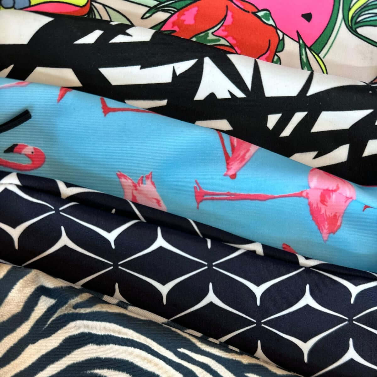 Fünf aufeinander gestapelte Stoffrollen, ideal für Hobby Horse-Projekte, jeweils mit unterschiedlichem Muster: bunte Früchte, schwarz-weißer abstrakter Text, Blau mit rosa Flamingos, Dunkelblau mit weißen geometrischen Formen und Zebrastreifen.