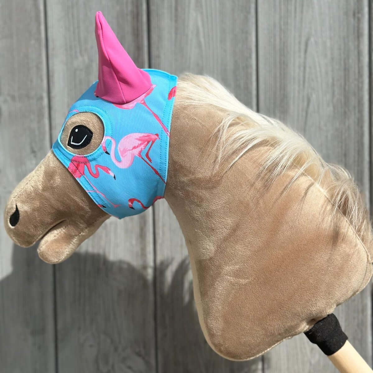 Ein Plüsch-Hobby Horse, ideal für Hobby Horsing, mit hellbraunem Fell und weißer Mähne, trägt eine blaue Kopfbedeckung mit rosa Flamingos und ein großes rosa Einhornhorn vor einem Holz-Hintergrund.