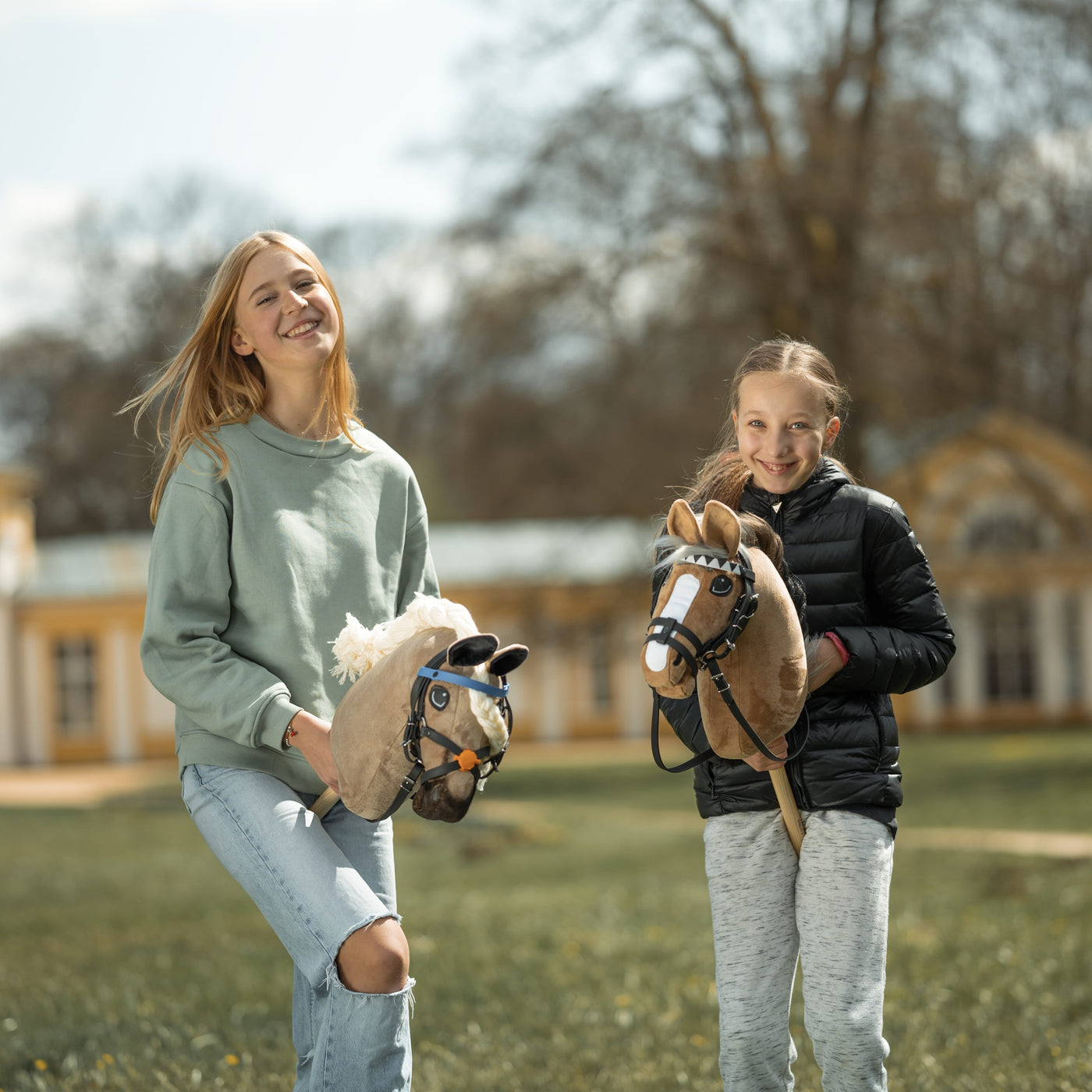 Zwei lächelnde Mädchen stehen draußen und halten ihre Hobby Horses. Eines trägt einen hellgrünen Pullover und zerrissene Jeans, das andere eine schwarze Jacke und graue Hose. Hobby Horsing bereitet ihnen Freude, während sie in der Nähe eines großen Gebäudes und mit Bäumen im Hintergrund posieren.