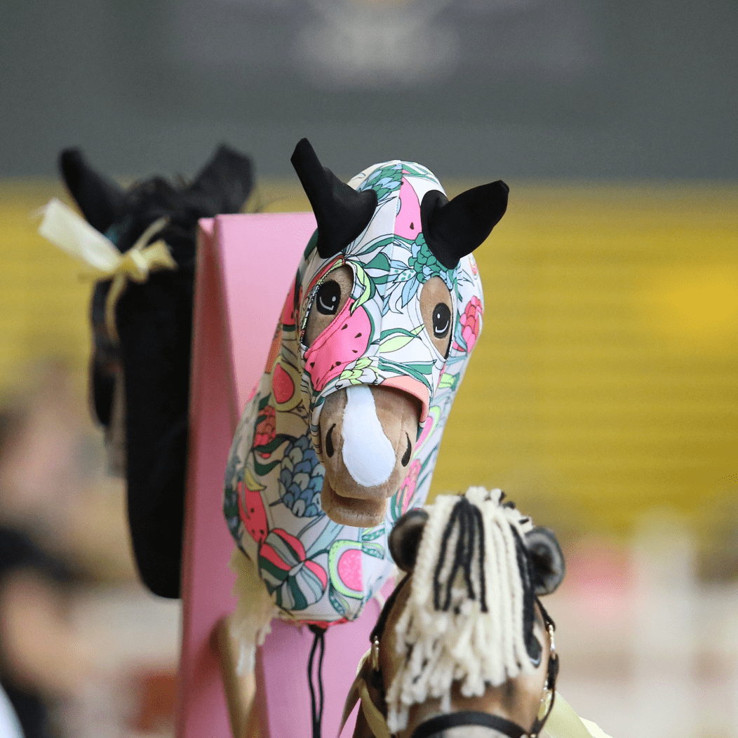 Eine Hobby Horsing-Szene zeigt ein Hobby Horse mit einer farbenfrohen Kopfbedeckung aus Stoff mit Blumenmuster und schwarzen Ohren im Fokus, umgeben von weiteren unscharfen Hobby Horses im Hintergrund.