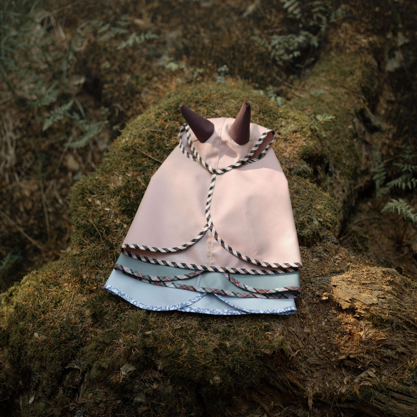 Ein kleiner rosa-blauer Stoffumhang mit schwarz abgesetzten Hörnern, perfekt für Hobby Horsing, liegt auf moosiger Baumrinde in einer Waldkulisse.