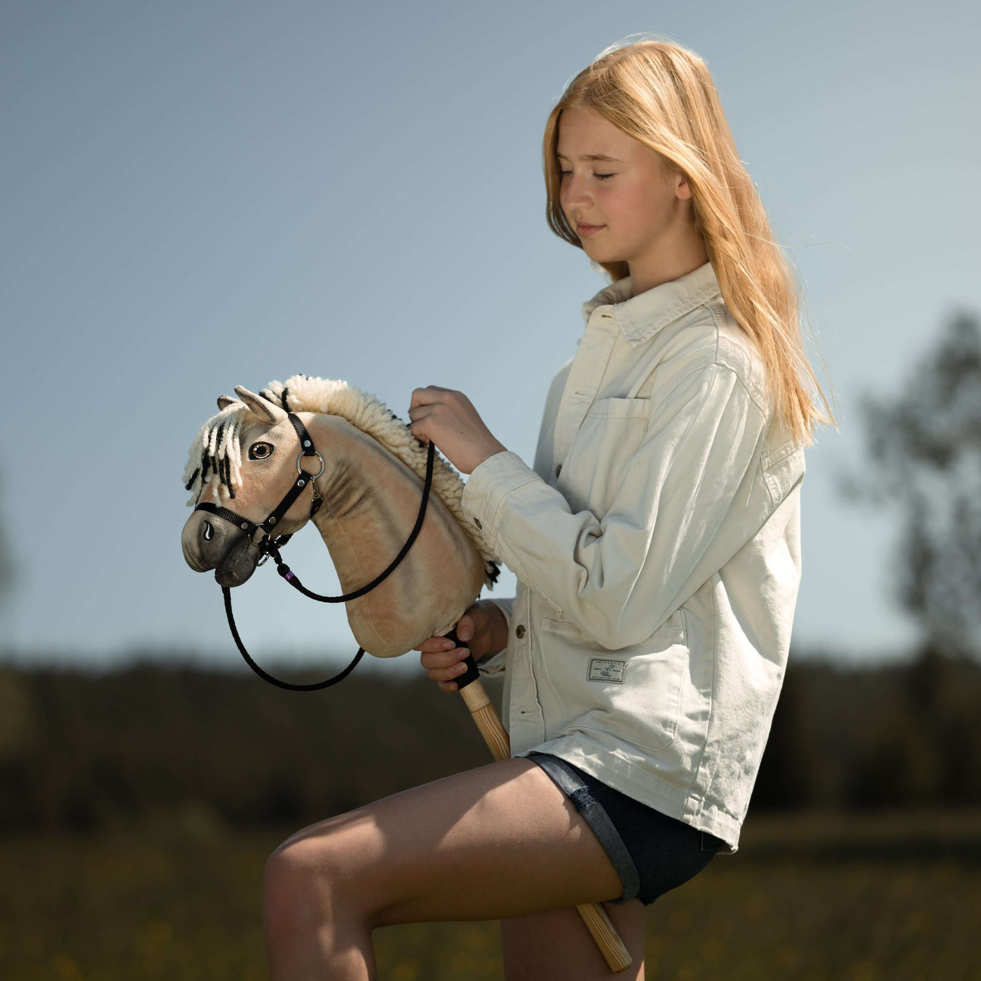 Ein junges Mädchen mit langen blonden Haaren, in einer leichten Jacke und Shorts, genießt an einem sonnigen Tag draußen Hobby Horsing und hält ihr Hobby Horse zwischen Bäumen und vor einem verschwommenen natürlichen Hintergrund.