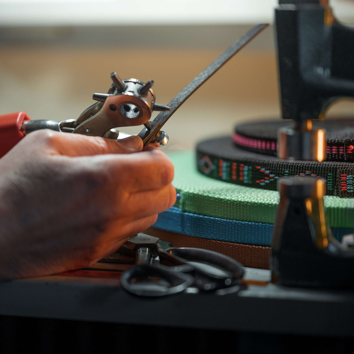 Nahaufnahme von Händen, die mit einem Locheisen an Leder arbeiten – perfekte Materialien zur Herstellung von Zaumzeug für Hobby Horse – mit Rollen aus buntem Gurtband, Nähwerkzeugen und einer Schere, die im Hintergrund auf der Werkbank verteilt liegen.