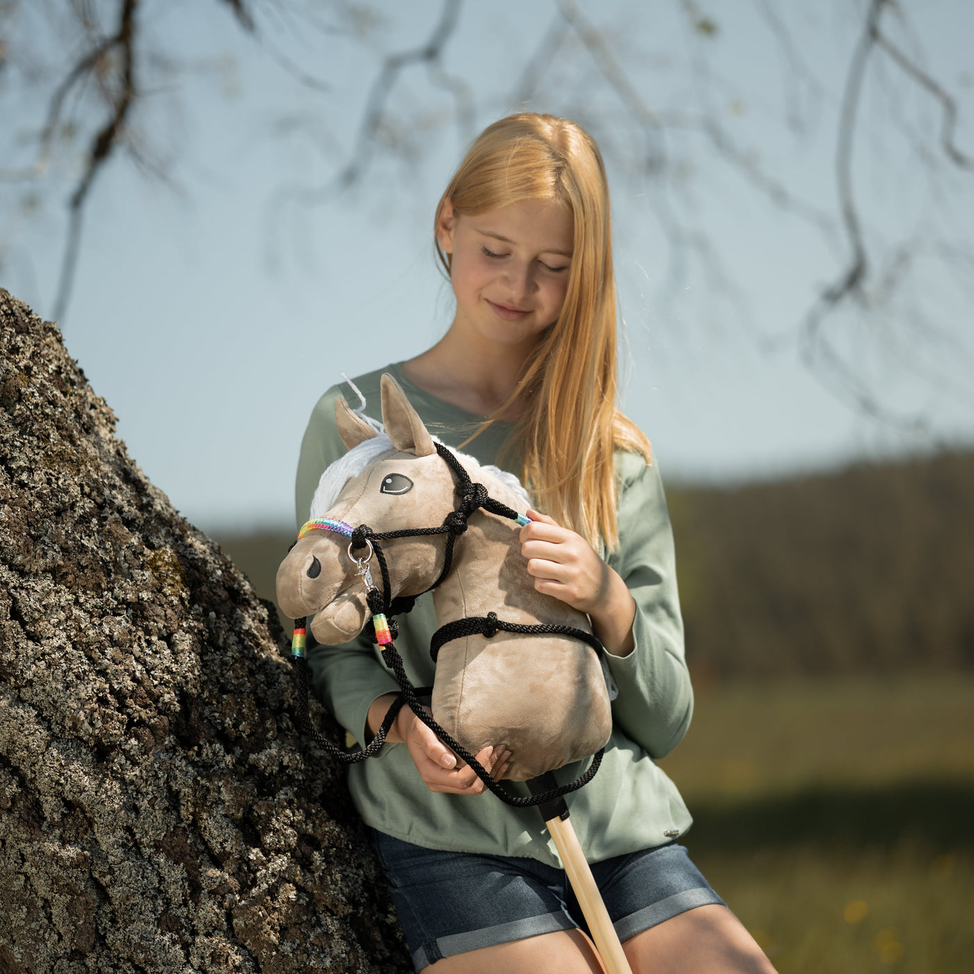 Ein junges Mädchen mit langen blonden Haaren, grünem Oberteil und Jeansshorts sitzt draußen an einem Baum und lächelt auf ihr Hobby Horse mit Zaumzeug und bunten Verzierungen hinab – es fängt den fröhlichen Geist von Hobby Horsing in einer natürlichen Umgebung ein.