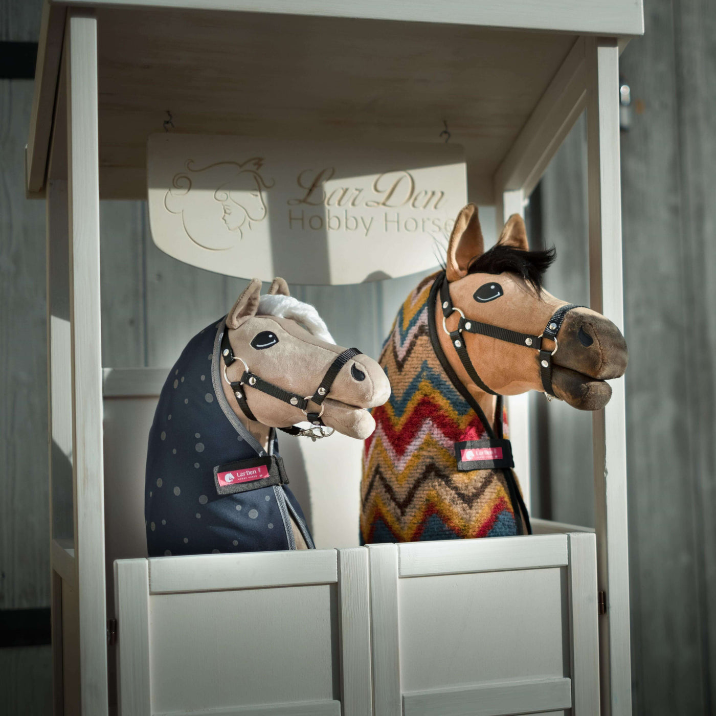 Zwei plüschige Hobby Horses mit Zaumzeug und Decken, eines mit blauer gepunkteter Decke und das andere mit buntem Zickzackmuster, sind nebeneinander in einem weißen Holzstand mit der Aufschrift LarsDen Hobby Horsing ausgestellt.
