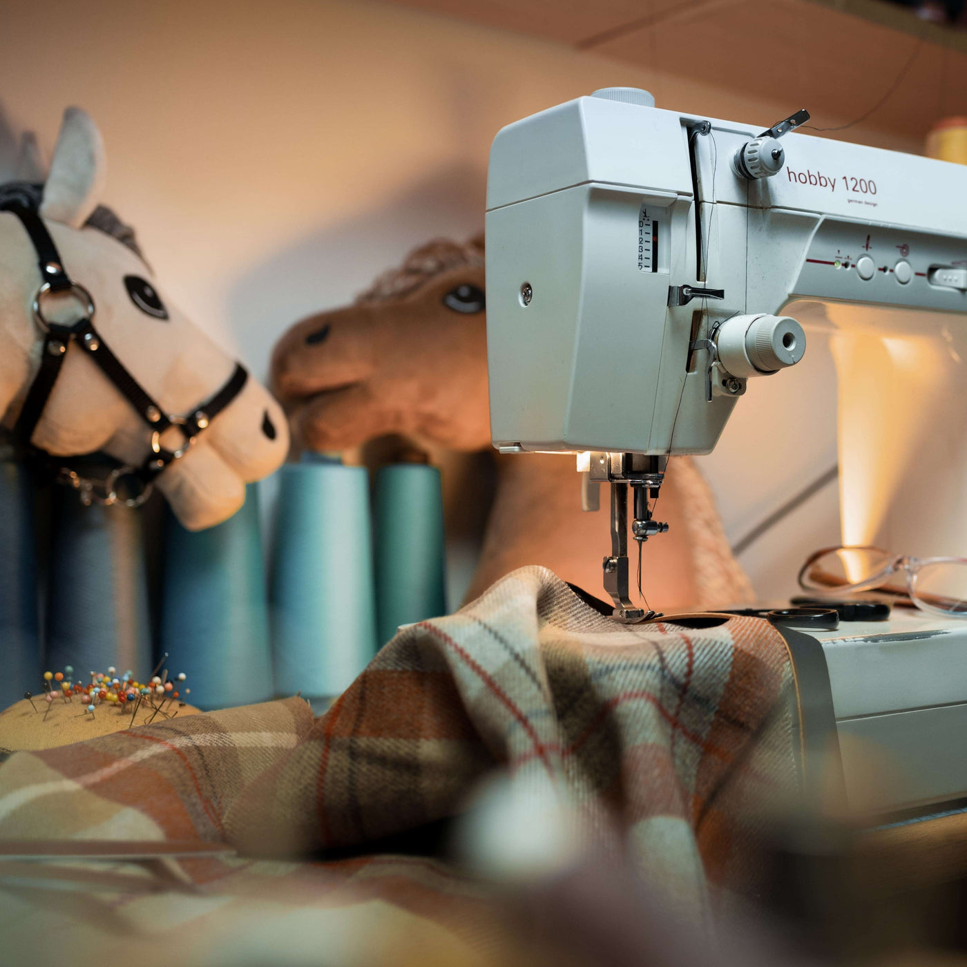 Eine Nähmaschine näht karierten Stoff, mit großen Spulen aus blauem Garn und zwei Plüsch-Hobby Horses im Hintergrund, und schafft eine gemütliche Bastelatmosphäre.