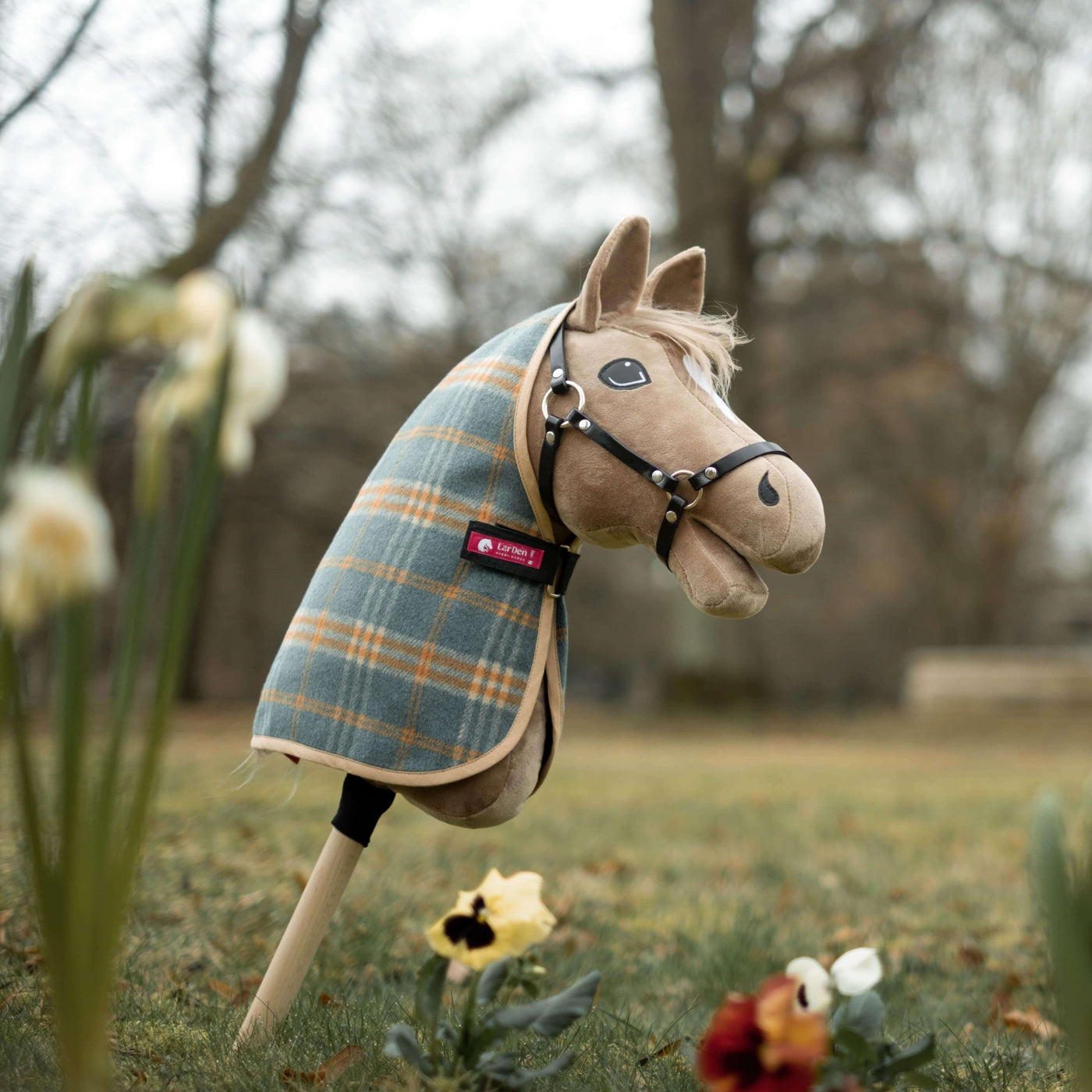 Ein Plüsch-Hobby Horse mit karierter Decke und Zaumzeug steht aufrecht auf einem Stock in einem grasigen Außenbereich, ideal als Bild für Hobby Horsing-Abenteuer zwischen Frühlingsblumen und Bäumen im Hintergrund.