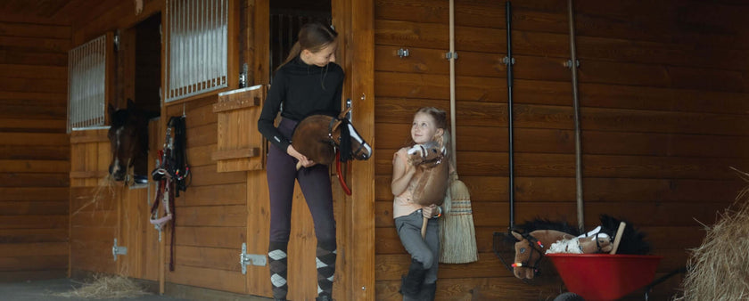 A woman and a young girl stand outside a wooden stable, each holding a hobby horse. Smiling at each other, they enjoy a moment of hobby horsing, surrounded by horse stalls, equipment, and a red wheelbarrow nearby.