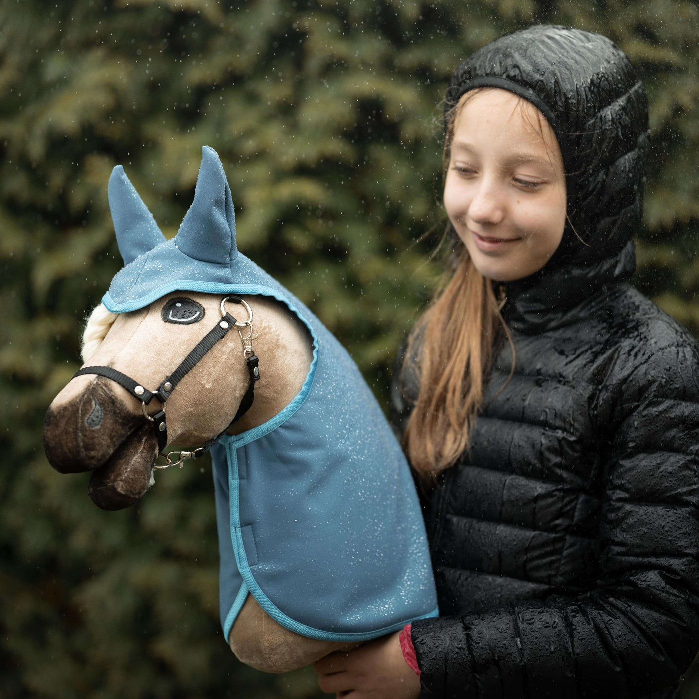 Ein lächelndes Mädchen in einer schwarzen Kapuzenjacke steht draußen im Regen und hält ihr geliebtes Hobby Horse Plüschtier in blauer Kleidung, mit unscharfem grünem Laub im Hintergrund – ein perfekter Moment für jeden Hobby Horsing Fan.
