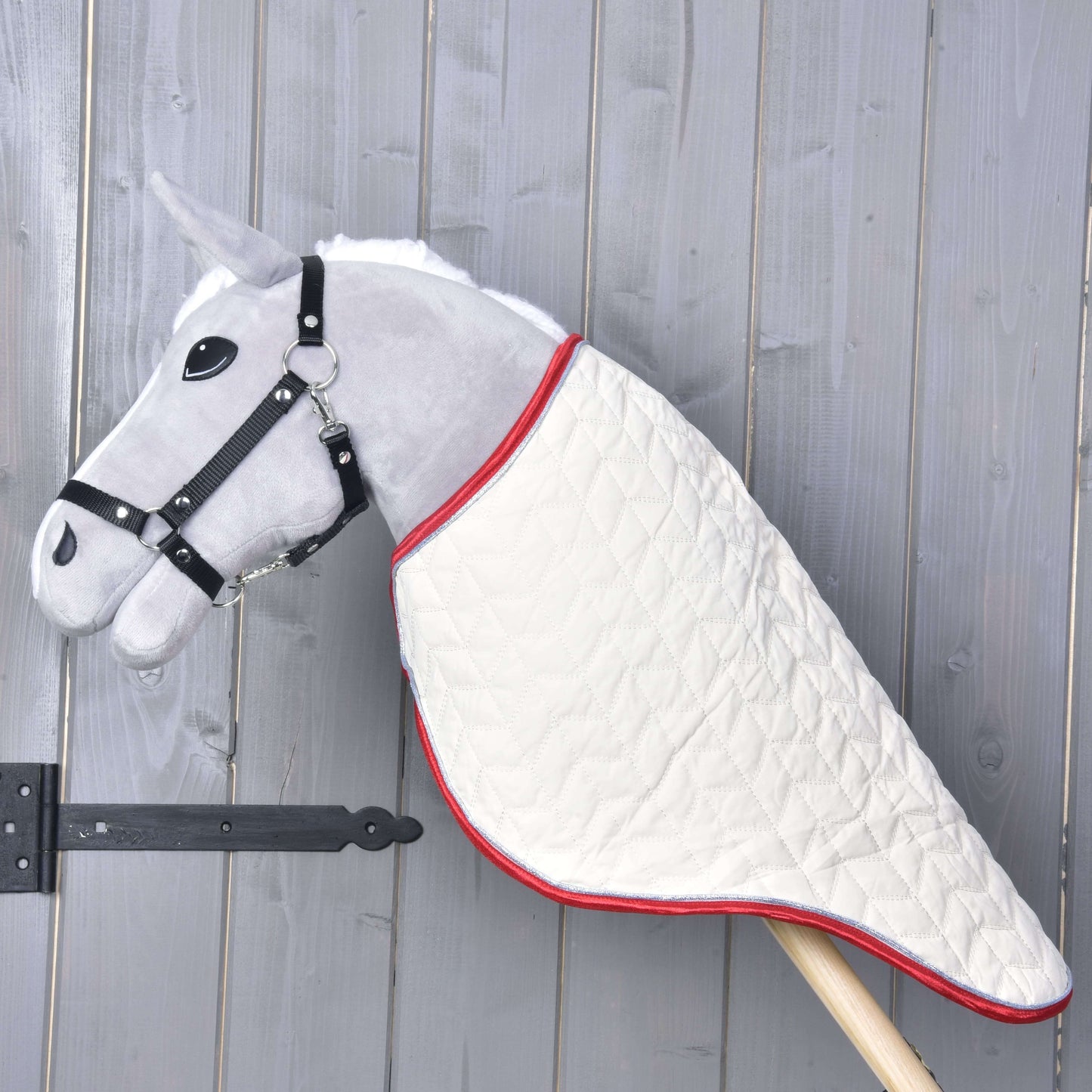 Die Hobby Horse LarDen Transportdecke Italy an einem grauen Plüsch-Hobby Horse mit schwarzem Zaumzeug und weißer gesteppter Decke auf einem Holzstab, gezeigt vor einer grauen Holzwand.