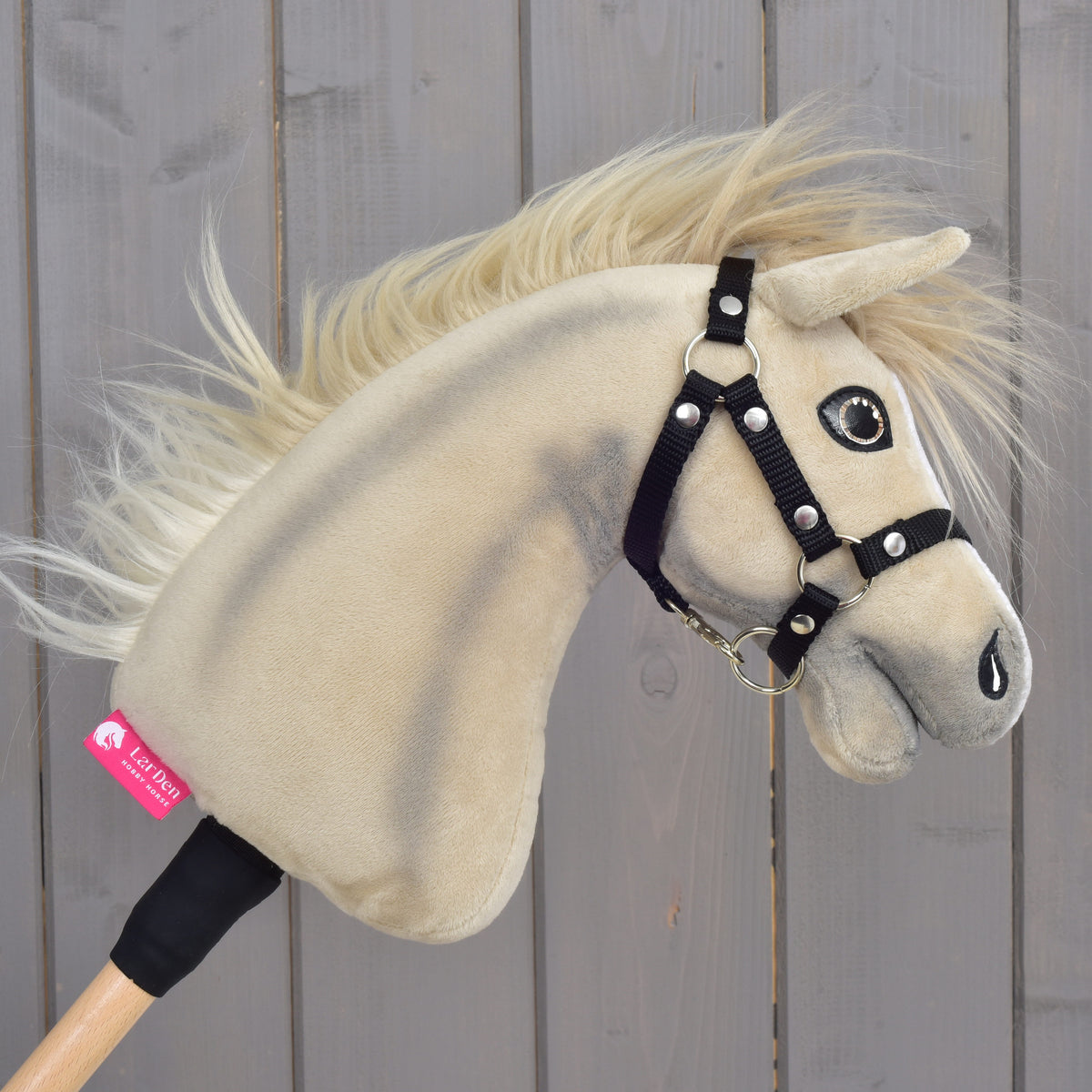 A close-up of the Hobby Horse LarDen “Hobby Horse Ivory Dream with Black Halter” in size XS, featuring a beige plush design with a black halter and a tag on its neck, set against a wooden plank background.
