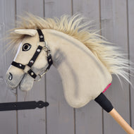 Hobby Horse Ivory Dream mit schwarzem Halfter – Größe XS von Hobby Horse LarDen in Größe XS, mit weichem elfenbeinfarbenem Fell und Holzstab, an eine graue Holzwand gelehnt.