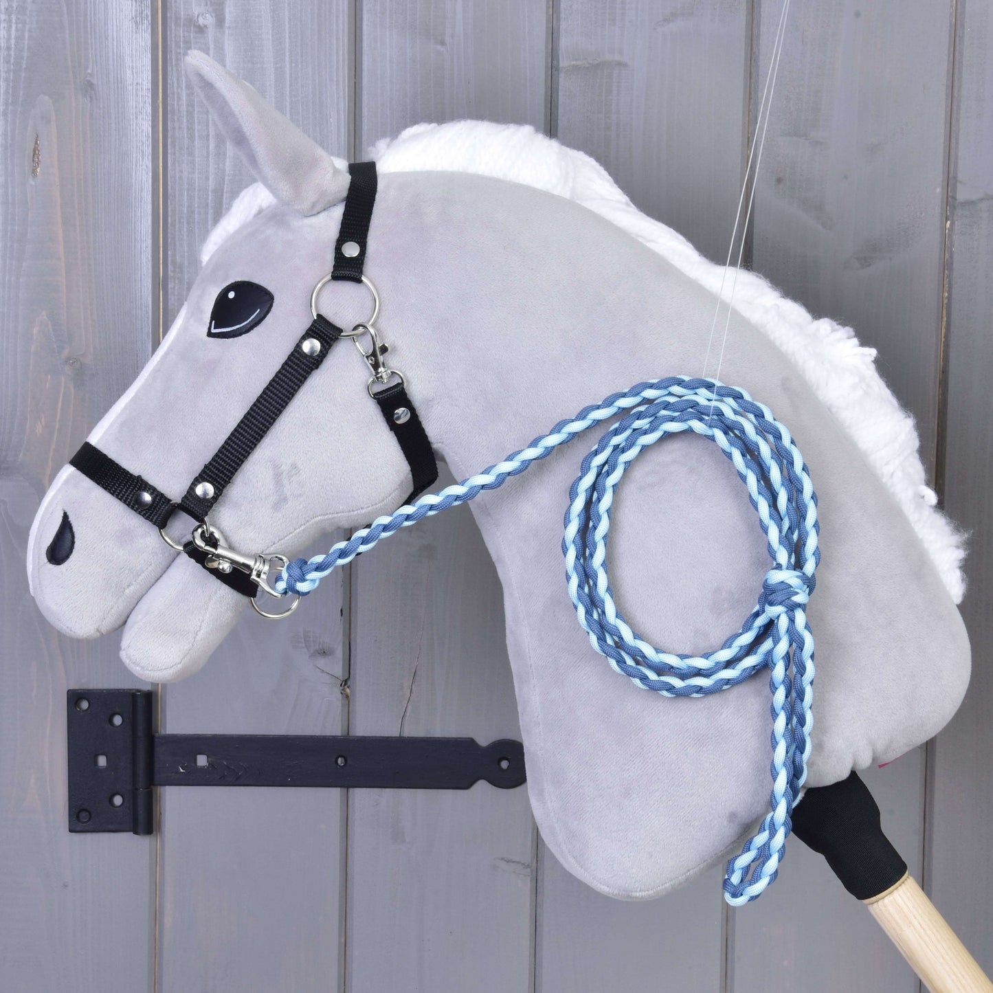 Das „Hobby Horse - Führstrick Knitted Double Blue“ von Hobby Horse LarDen zeigt ein graues Plüschpferd mit schwarzem Zaumzeug für Hobby Horse und einem handgestrickten blauen Führstrick mit hochwertigem Karabiner, vor einer Holzwand.