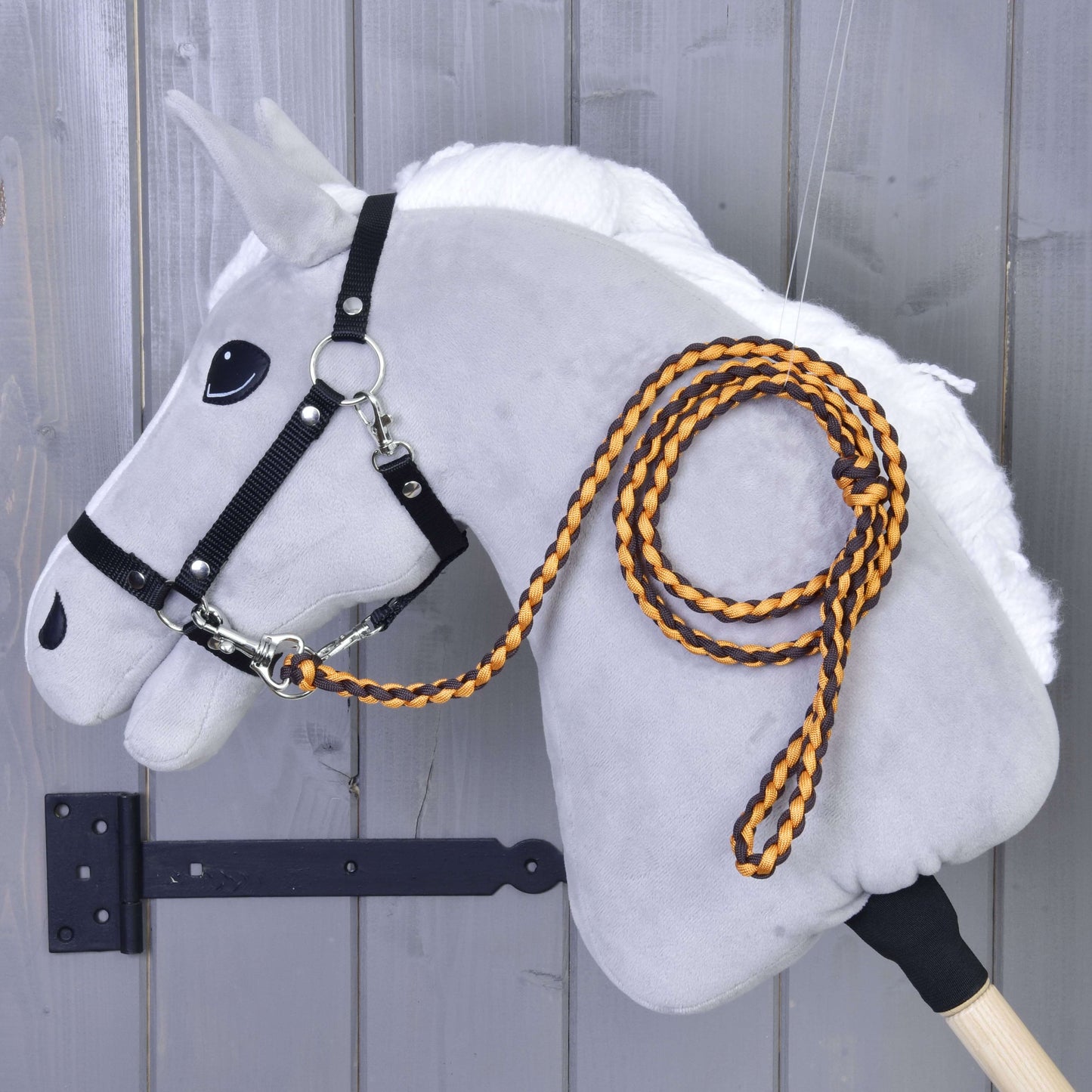 Das „Hobby Horse - Führstrick Knitted Pomello“ von Hobby Horse LarDen zeigt ein graues Plüsch-Hobby Horse mit weißer Mähne, schwarzem Zaumzeug für Hobby Horse und einem orange-schwarzen gestrickten Führstrick mit Karabiner, montiert an einer Holzwand.