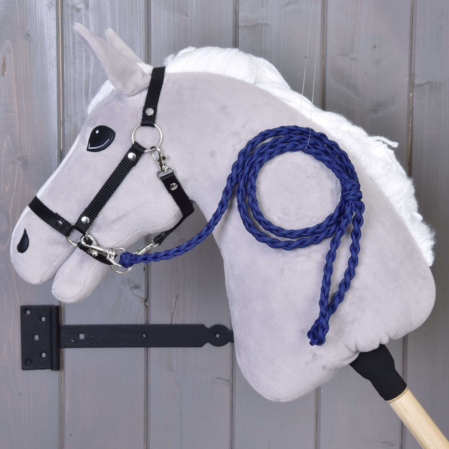 Ein graues Plüsch-Hobby Horse - Führstrick Knitted Royal Blue von Hobby Horse LarDen mit weißer Mähne, schwarzem Zaumzeug für Hobby Horse und royalblauem handgestricktem Führstrick, an einer Holzwand montiert, wobei der Stab mit einer schwarzen Metallhalterung befestigt ist.