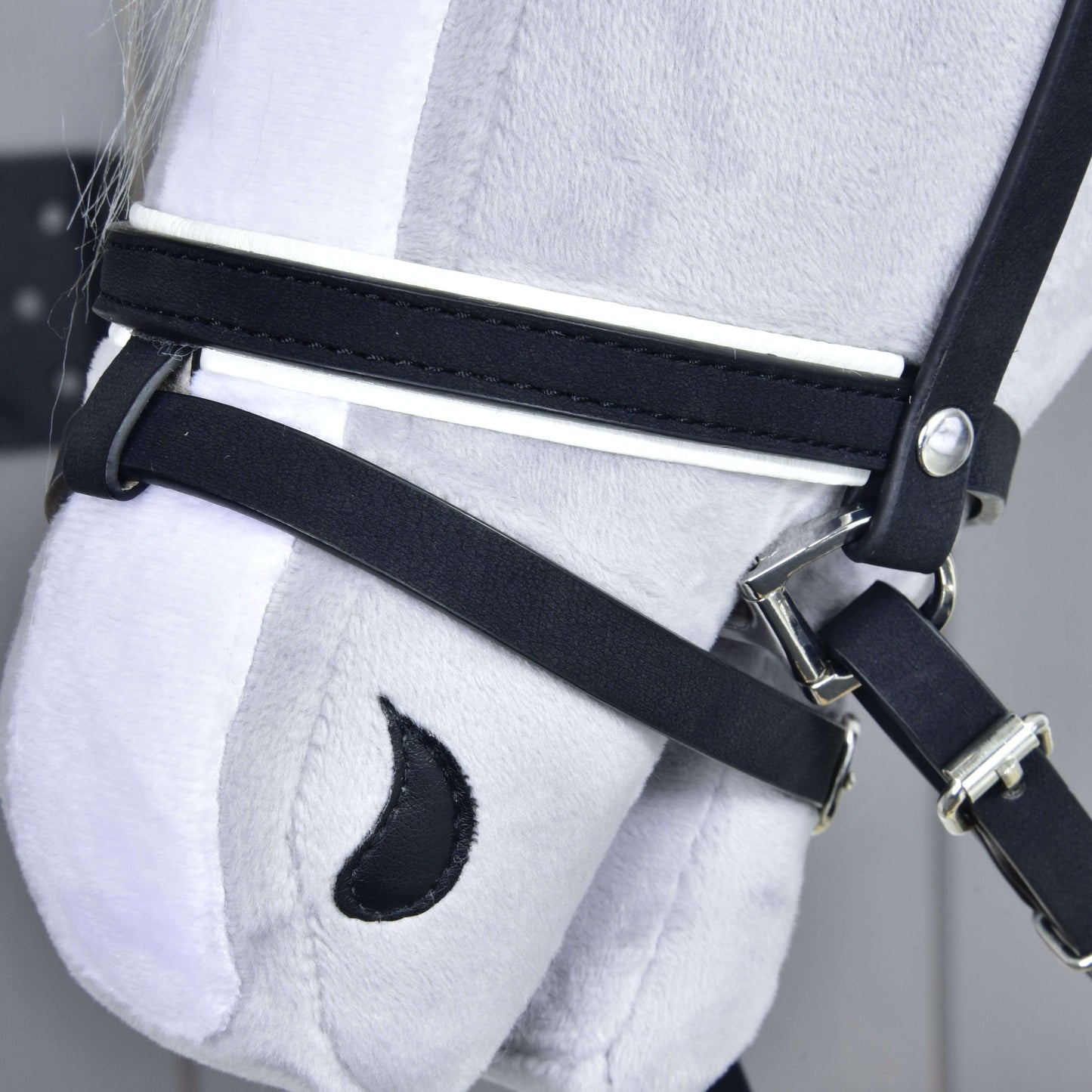 Nahaufnahme eines grauen Plüschpferdekopfs von Hobby Horse LarDen mit Komplette De Luxe Trense mit 2 Stirnbänder. Gestickte Details wie ein schwarzes halbmondförmiges Nasenloch heben sich vor hellem Hintergrund ab.