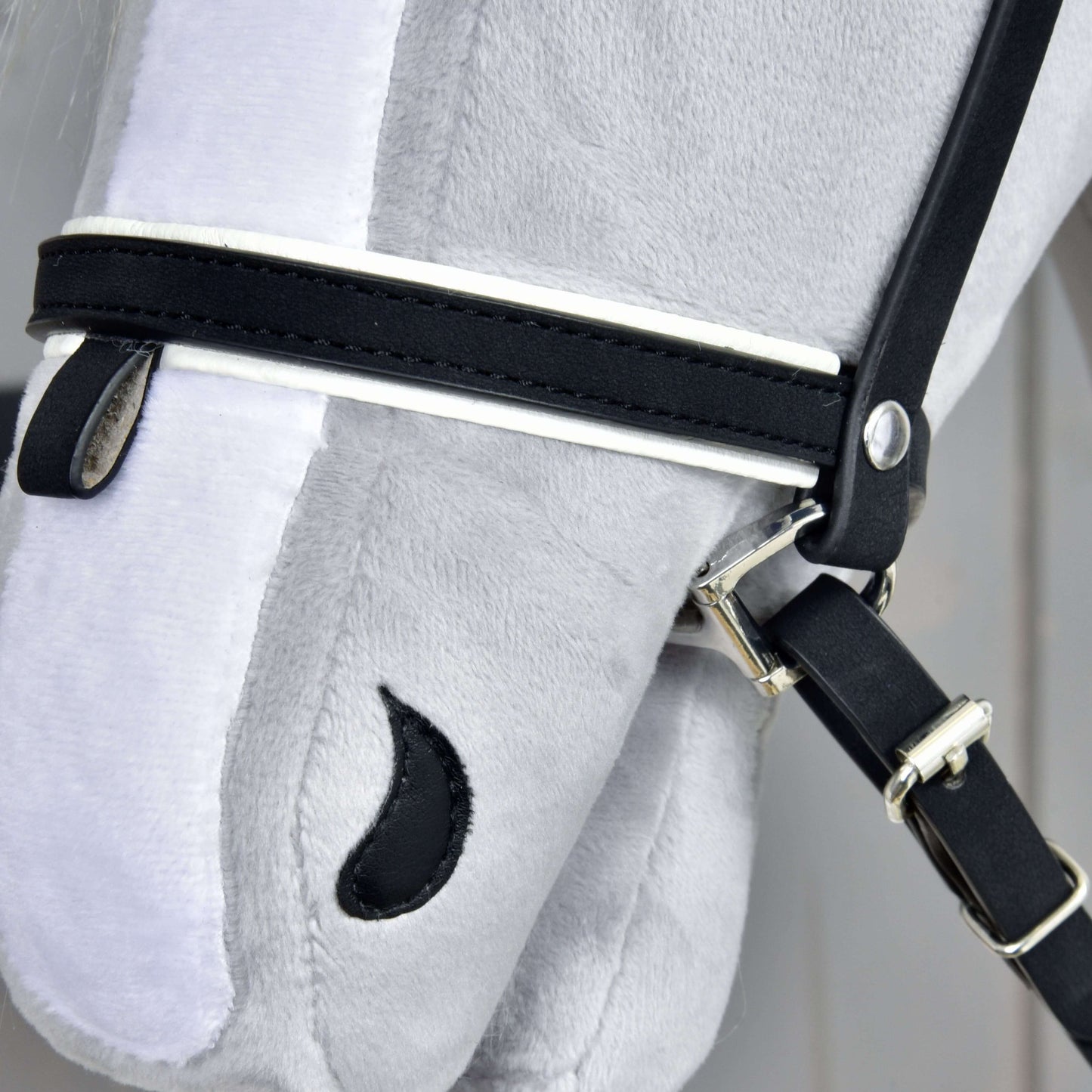 Komplette De Luxe Trense mit 2 Stirnbänder von Hobby Horse LarDen, Nahaufnahme des Kopfes eines grauen Plüsch-Hobby Horse, mit 2 abnehmbaren Stirnbändern, kombiniertem Nasenriemen, Metallschnallen und einem auf weichen Stoff gestickten schwarzen Auge.