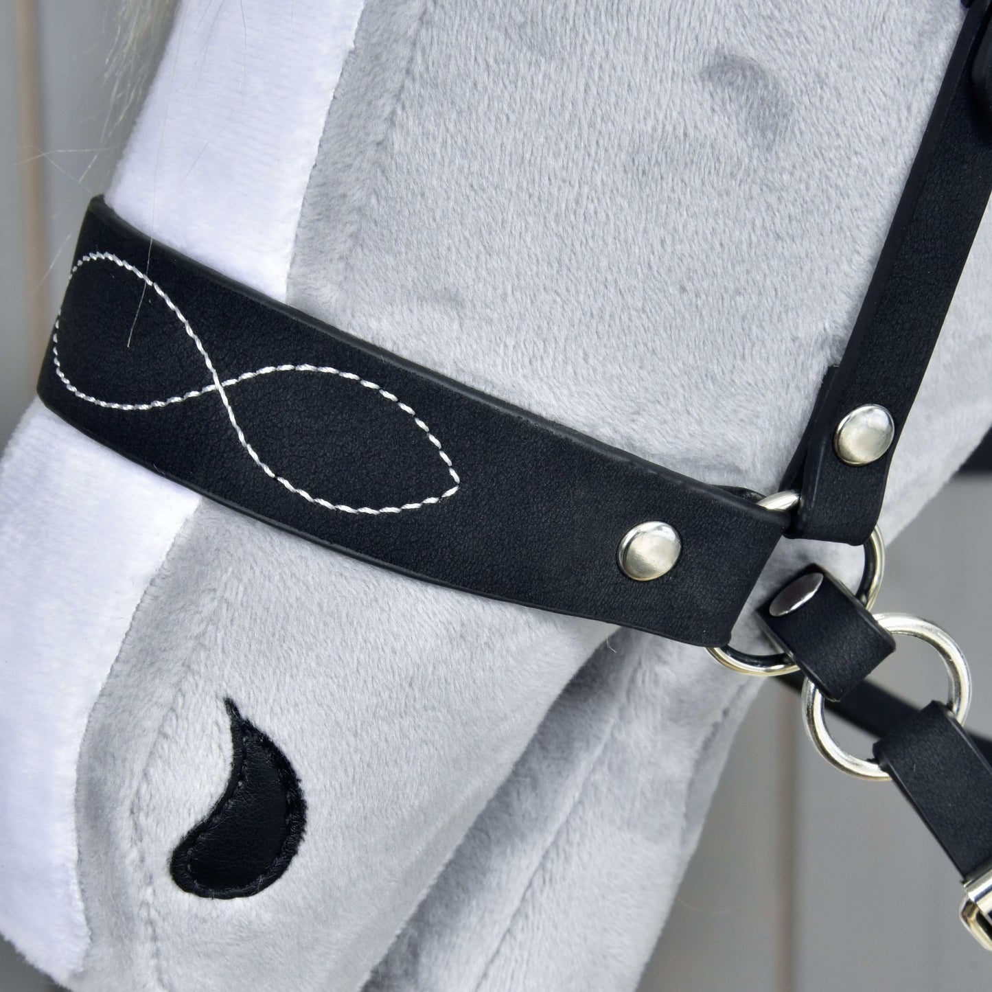 Nahaufnahme eines grauen Plüsch-Hobby Horse-Kopfs mit Hobby Horse LarDen Komplette gebisslose Trense, verziert, mit weißer Stickerei in Form eines Unendlichkeitszeichens und einem schwarzen tropfenförmigen Einsatz nahe dem Auge.