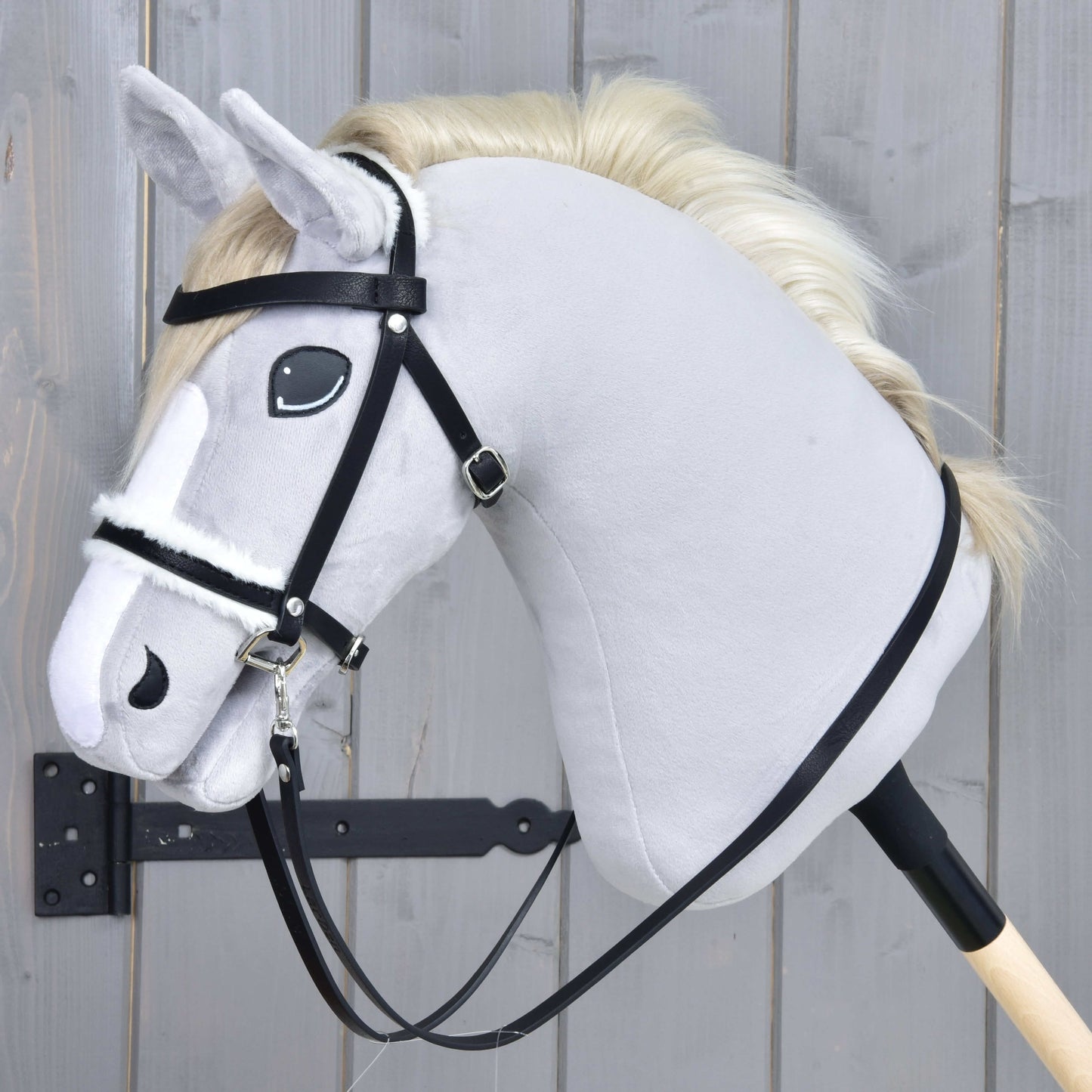 Hobby Horse LarDen Komplette Trense mit Fell an einem hellgrauen Hobby Horse mit cremefarbener Mähne, schwarzer Trense aus Kunstleder und Zügeln auf einem Holzstab, vor grauem Holzhintergrund.